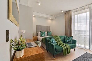 Ein modernes Schlafzimmer mit einem Bett, einem grünen Sofa, einem Schreibtisch mit Laptop, einer Topfpflanze und einem großen Fenster mit Vorhängen.