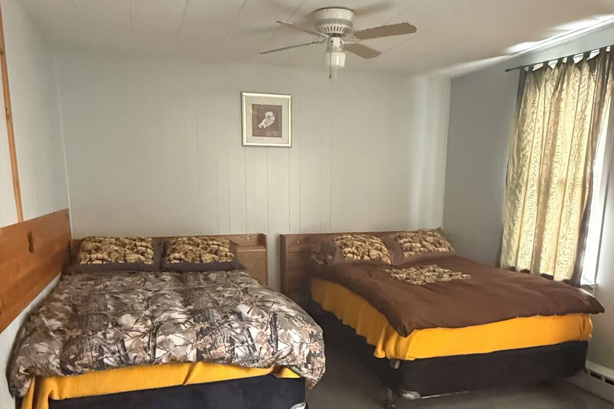 A room with two beds, a ceiling fan, a framed picture on the wall, and a window with curtains.
