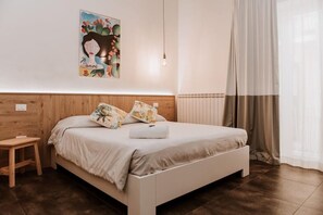 Un dormitorio con una cama, una mesita de noche de madera, una lámpara de pared, un radiador y una ventana con cortinas.