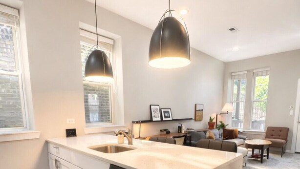 A modern kitchen with a dishwasher, a sink, and a large island. There is a trash can and a dining area with a table and chairs. - Flexhome West Town Spacious 2BR Duplex · Sleeps 8 (Chicago)