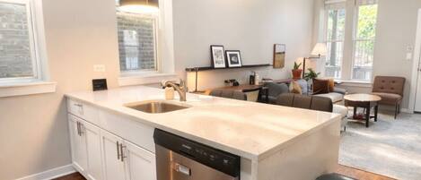A modern kitchen with a dishwasher, a sink, and a large island. There is a trash can and a dining area with a table and chairs.