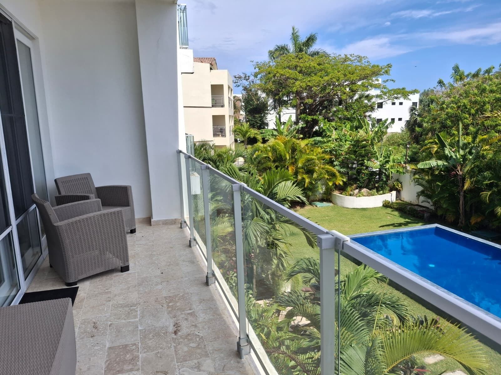A balcony with wicker furniture, a glass railing, and a view of a swimming pool and greenery.