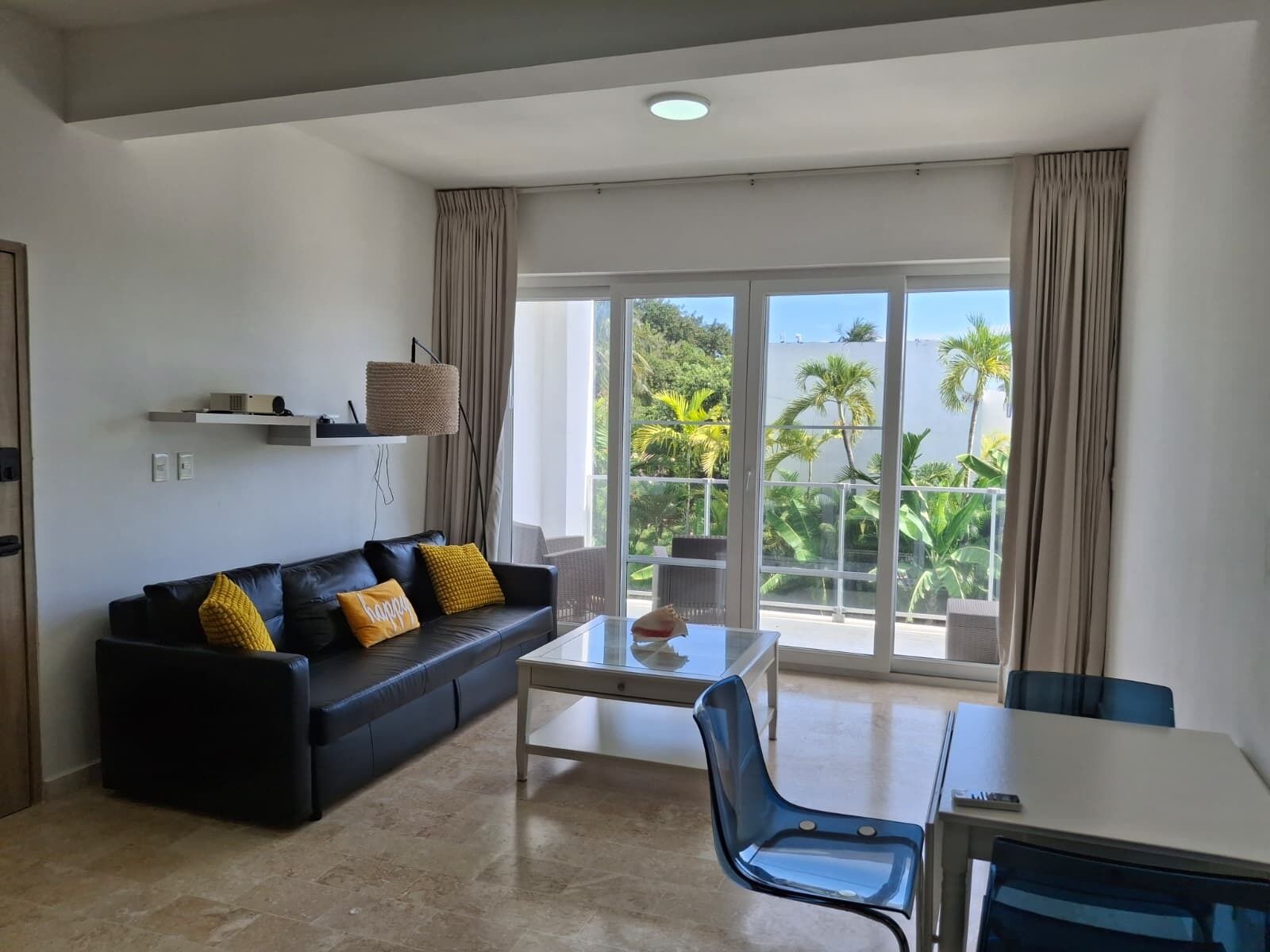 Un salón moderno con un sofá negro, una mesa de centro de vidrio, una mesa de comedor y vistas a palmeras a través de amplios ventanales.