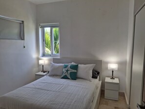 A bedroom with a bed, two bedside tables, a window with a view of greenery, and a wall-mounted light fixture. - 2 Bedroom, Pet friendly, Surf/ Kite, Encuentro Gem (Cabarete)