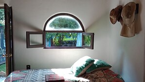 Un lit avec une literie à motifs, une fenêtre donnant sur un paysage verdoyant et un luminaire en fibres tressées.