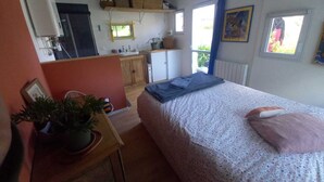 A bedroom with a bed, a desk with a plant, a kitchen area, and a window with curtains. - Dependent room at the bottom of the garden (Perros-Guirec)