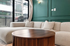 Una sala de estar moderna con un sofá seccional, una mesa de centro redonda de madera y vistas a un paisaje urbano a través de grandes ventanales.