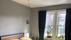 Ein Schlafzimmer mit Bett, Nachttisch, Deckenventilator, einem Fenster mit Vorhängen und Blick auf Gebäude.