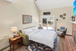 A bedroom with a bed, a nightstand, a lamp, a chair, a fireplace, and a window with a view of the outdoors.