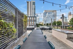 Un área de comedor al aire libre con una larga mesa y sillas, una valla de madera con plantas trepadoras y un horizonte urbano de fondo.