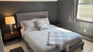 Une chambre à coucher avec un lit, des tables de chevet, une lampe, une fenêtre et une vue sur la verdure à l’extérieur.