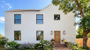 A two-story white brick house with a front door, windows, and a small front yard with plants.