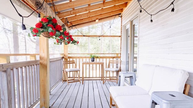 Un terrazzo in legno con un divano da esterno bianco, un tavolo con sedie e fiori rossi pendenti.