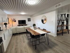 A modern kitchen with a built-in oven, a dining area with a wooden table and chairs, a white sofa, and a TV mounted on the wall. - Iron & Oak Suite Minutes from Silverstar Skii Resort  Lakes, Hiking &Restaurant (Vernon)