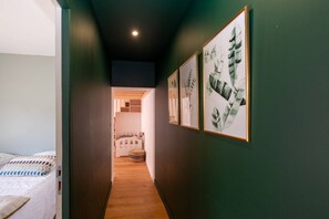 Ein schmaler Flur mit einem Bett auf der linken Seite, einer Tür, die zu einem Zimmer mit einem Bett führt, und gerahmten Kunstwerken an den Wänden.