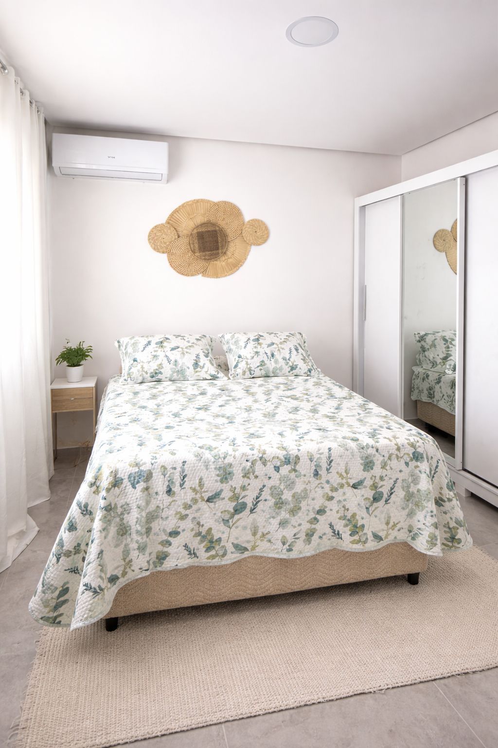 A bedroom with a bed, a bedside table, a wall-mounted air conditioner, a mirror, and a decorative wall hanging.