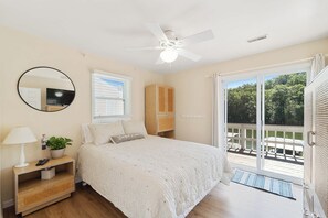 Ein Schlafzimmer mit Bett, Nachttisch, Spiegel, Deckenventilator, Fenster und einer Glastür, die auf eine Terrassenfläche führt.