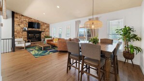 A spacious living area with a fireplace, a dining table with chairs, a sofa, and a television mounted on a stone wall. - Lake Front |Private Dock |Blue Bungalow|3bed|2bth (Linn Creek)