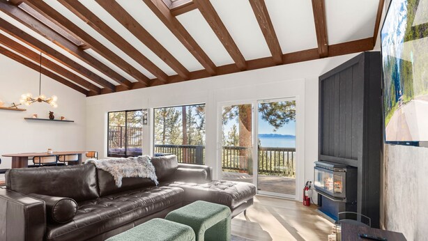 A modern living room with a leather sofa, ottomans, a fireplace, and large windows offering a view of the outdoors. - Lake View 3BR/2BA in Incline Village, Fireplace (Incline Village)