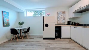 A modern kitchen with white cabinets, a black refrigerator, a dining table with two chairs, and a window providing natural light. - Perfect 1-bedroom basement with AC in pleasant Burnaby (Burnaby)