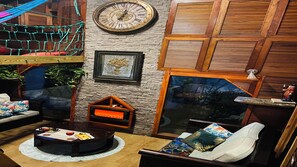 A cozy room with a stone wall, a wooden ceiling, a large clock, a fireplace, and a window with a view of trees.