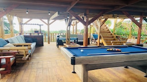 Una terraza de madera con mesa de billar, sillones de descanso y una escalera que sube a un nivel superior.