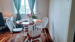 A dining area with a round wooden table, white chairs, a vase with flowers, and a window with curtains.
