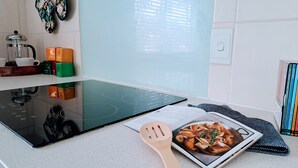 A kitchen with a black induction cooktop, a wooden spatula, a cookbook, and a window with blinds.