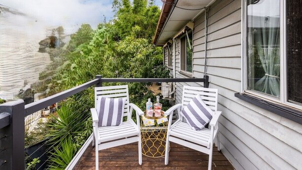 A wooden deck with two white chairs and a small table, surrounded by greenery and a house with white siding.