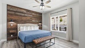 Een slaapkamer met een houten wand, een bed met een blauwe sprei, een bankje, een plafondventilator en een raam met gordijnen.