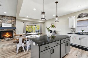 A modern kitchen with a stone fireplace, a dining area with white chairs, and a kitchen island with cabinets. - Jubilee Retreat+7 King Suites+Sleeps 24+Hot Tub+Table Rock Lake+Game Room (Branson)
