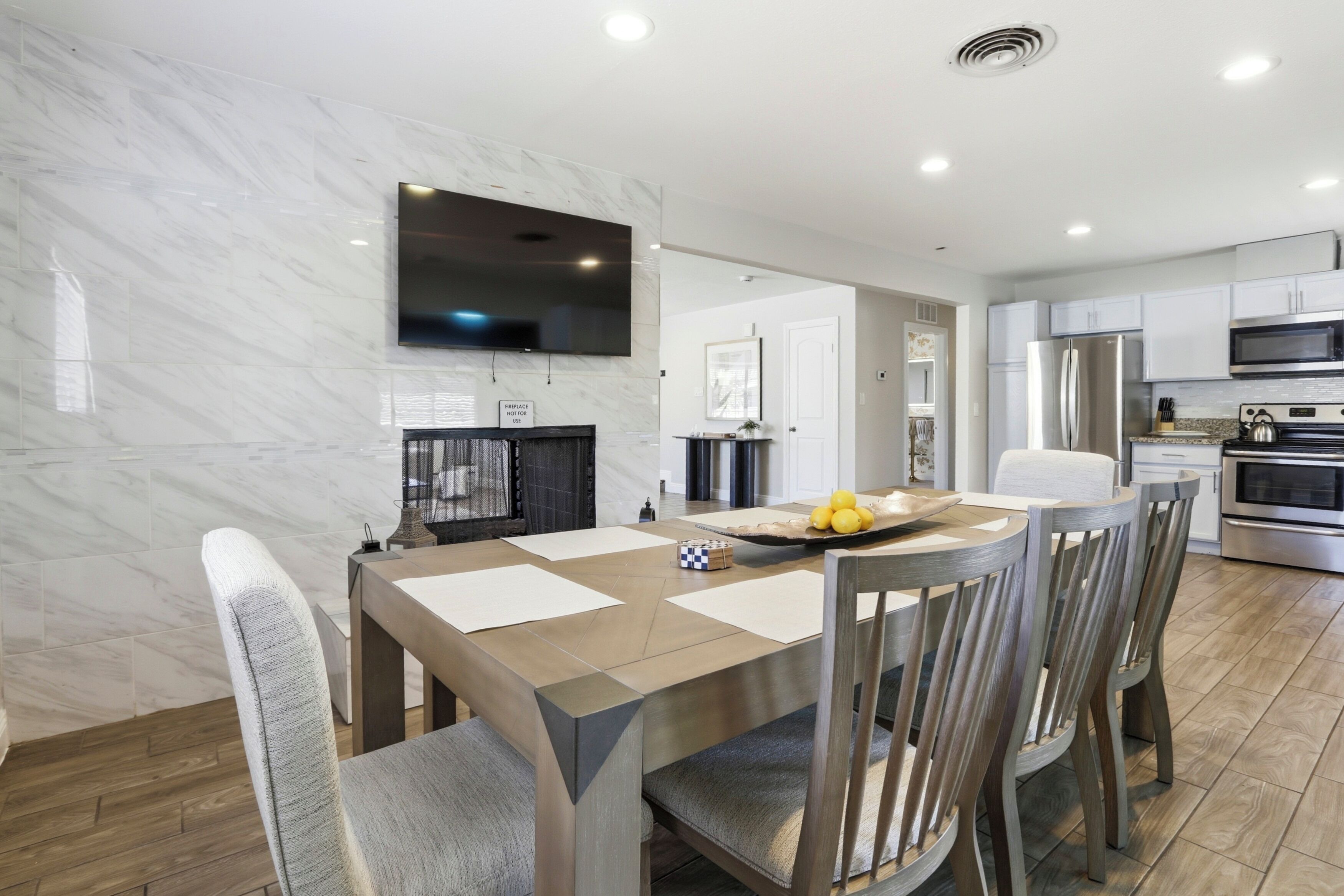 A modern kitchen with a flat-screen TV, stainless steel appliances, and a dining area with a marble backsplash and wooden table.