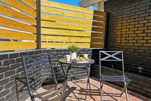 Eine Terrasse mit zwei Metallstühlen, einem kleinen runden Tisch mit einer Pflanze und einem Teller Essen sowie einer Ziegelwand.
