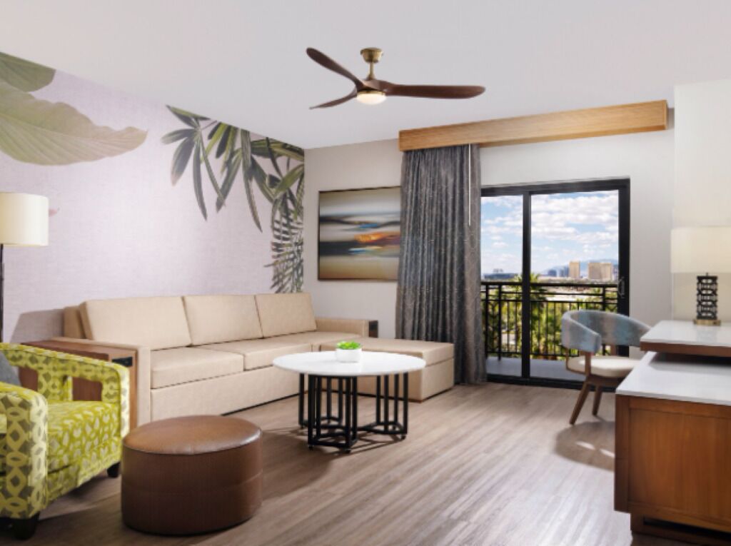 A modern hotel room with a sofa, a round coffee table, a chair, a desk, a ceiling fan, a large window with a view, and a painting on the wall.