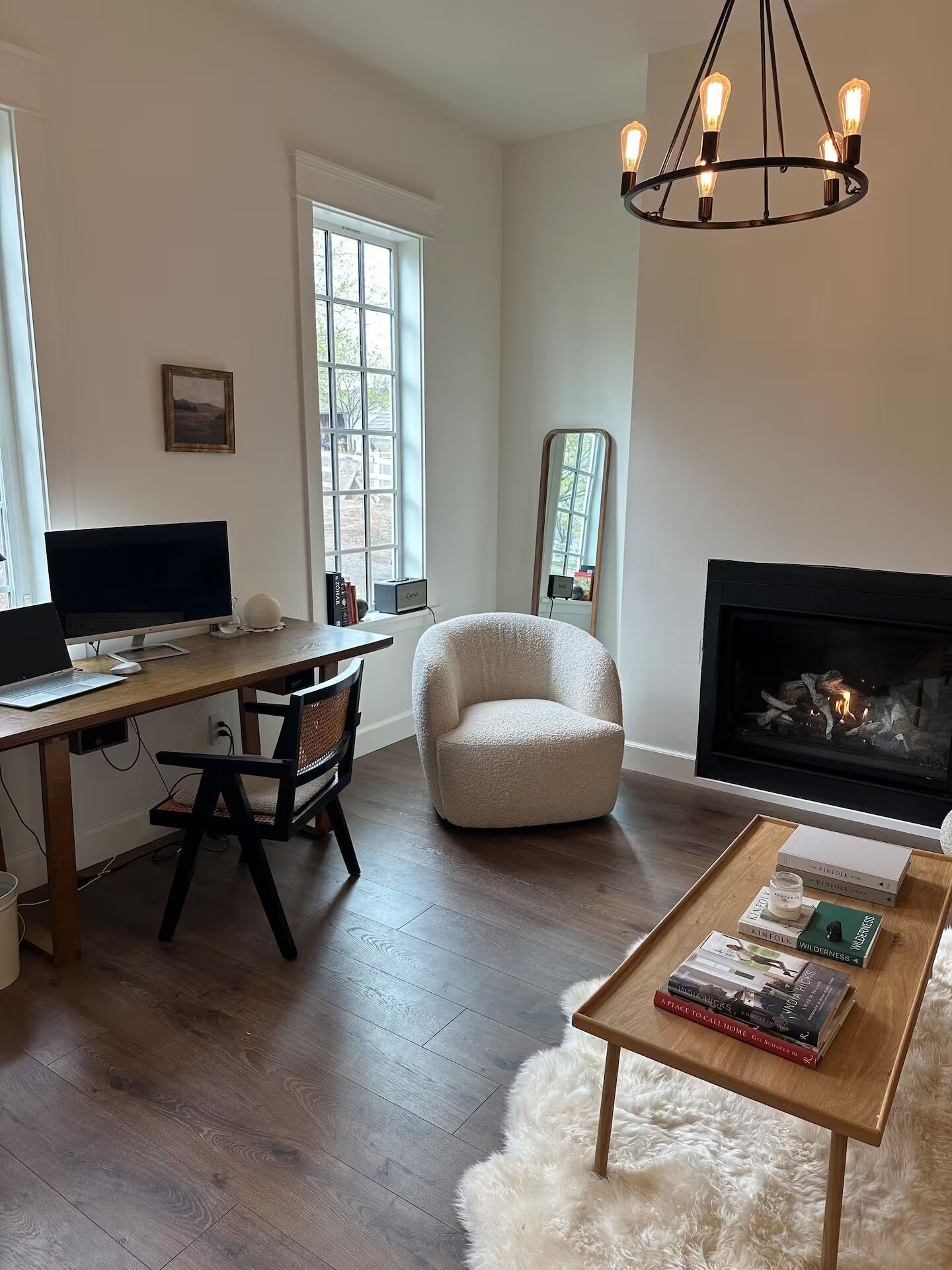 A modern living room with a fireplace, a desk with a computer, a comfortable armchair, and a coffee table with books.