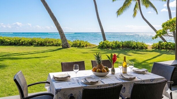 A table set outdoors with a view of the ocean, surrounded by greenery and palm trees. - Oceanfront Two-Bedroom Condo, Steps from the Sand and Pool (Kapaʻa)