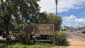A sign with palm tree and text, surrounded by greenery and a road. - Oceanfront Two-Bedroom Condo, Steps from the Sand and Pool (Kapaʻa)