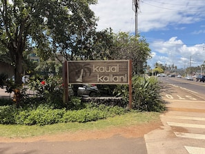 A sign with palm tree and text, surrounded by greenery and a road. - Oceanfront Two-Bedroom Condo, Steps from the Sand and Pool (Kapaʻa)