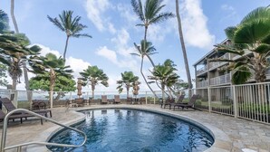 A pool area with lounge chairs and palm trees, surrounded by a stone deck and a white fence. - Oceanfront Two-Bedroom Condo, Steps from the Sand and Pool (Kapaʻa)