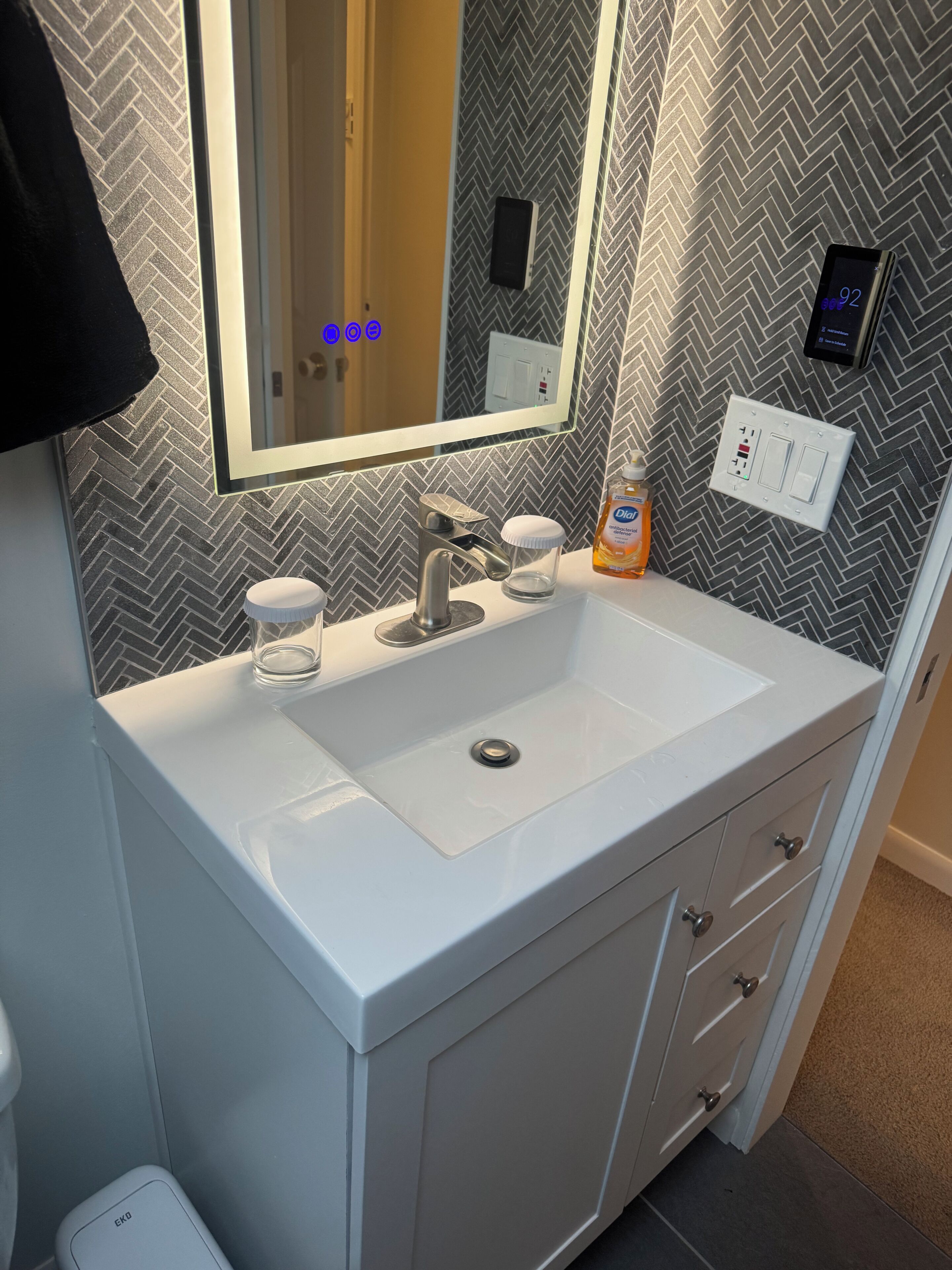 A bathroom with a white sink, a mirror, a herringbone-patterned wall, a soap dispenser, and a digital thermostat.