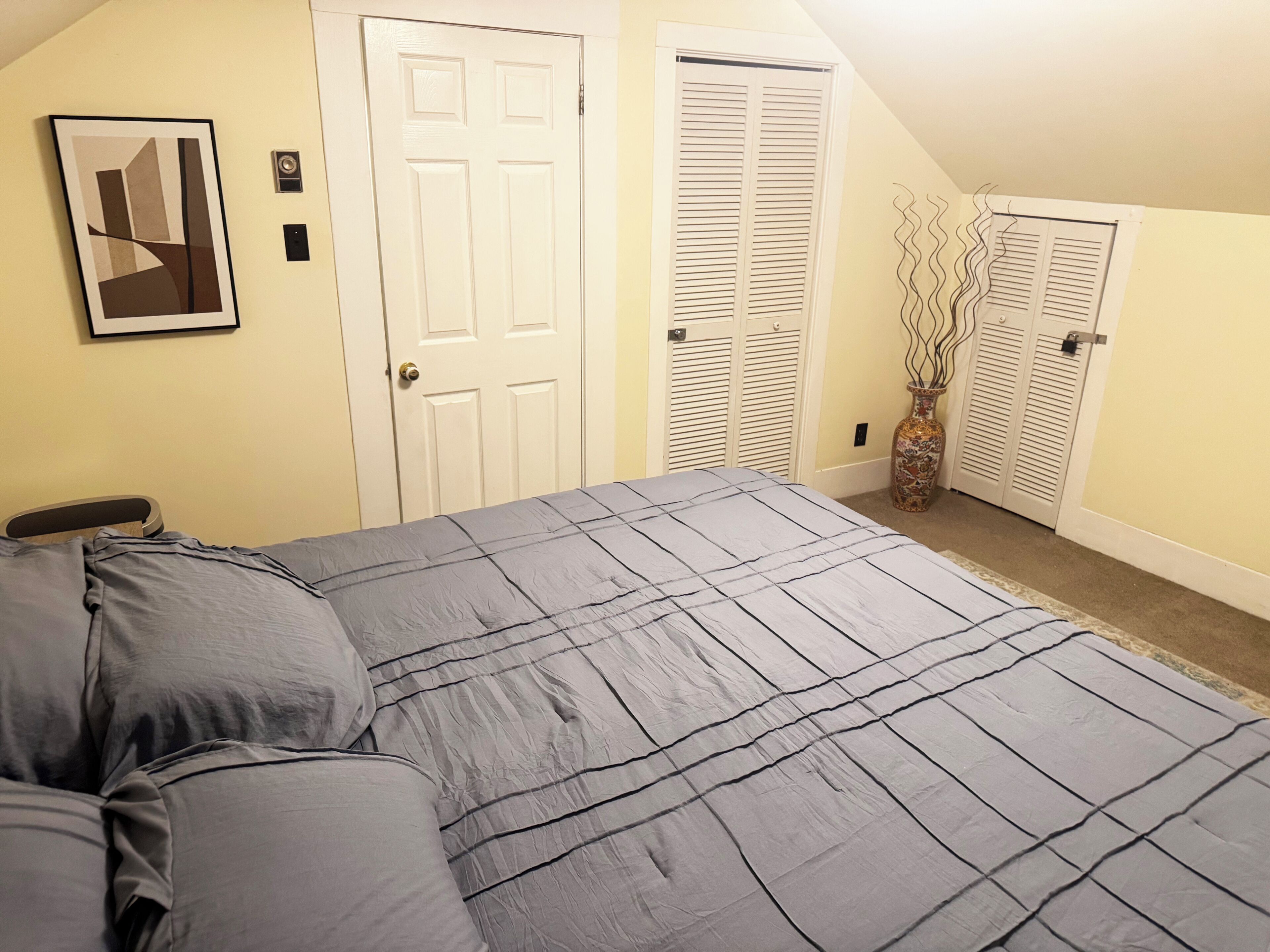 Un dormitorio con una cama, dos puertas blancas, un florero y un cuadro en la pared.