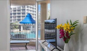 Un balcón con vista a la piscina, una sombrilla azul y un jarrón con flores.