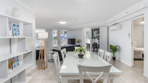 A modern dining area with a glass table, white chairs, and a white shelving unit. - Moroccan Surfers - 3BR Apartment (Surfers Paradise)