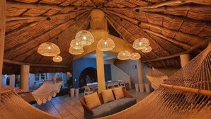A thatched-roof structure with hanging woven light fixtures, a hammock, and a dining area with chairs and a table.