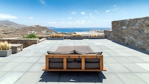 A rooftop terrace with a wooden bench, stone walls, and a view of the sea and distant hills.