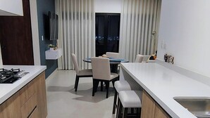 A modern kitchen with a central island, a dining area with chairs, a TV mounted on the wall, and a window with curtains.