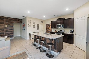A modern kitchen with a central island, dark cabinetry, stainless steel appliances, and a tiled floor. - 6BR Themed Villa w/ Game Room+ Pool & Spa Solterra (Davenport)