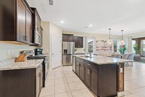 A modern kitchen with dark cabinetry, stainless steel appliances, and a large island with a sink. - 6BR Themed Villa w/ Game Room+ Pool & Spa Solterra (Davenport)