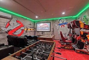 A room with a foosball table, a TV screen displaying a movie, and a wall with a Ghostbusters-themed mural. - 6BR Themed Villa w/ Game Room+ Pool & Spa Solterra (Davenport)