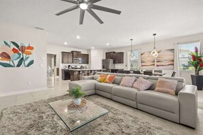Una sala de estar moderna con un sofá, una mesa de centro de cristal, un ventilador de techo y una cocina al fondo.